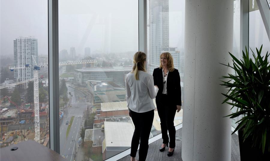 Two women stand facing each other in front of a floor to ceiling window that looks out over a city.